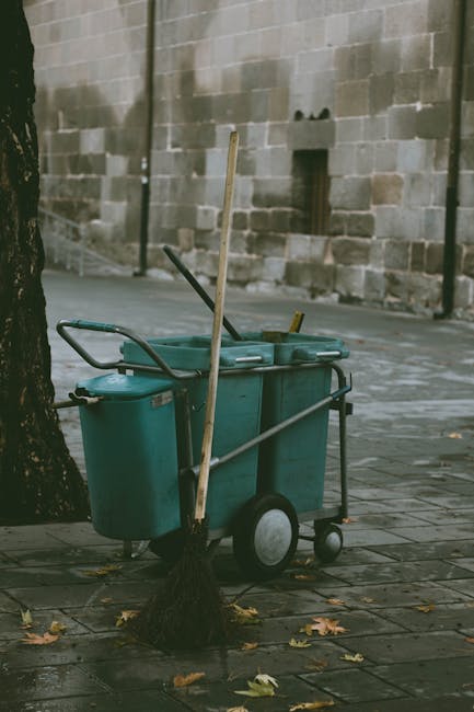 A cleaning trolley with multiple compartments and large wheels, positioned on a paved sidewalk near a tree with fallen autumn leaves. The trolley contains cleaning tools such as mop handles and a broo