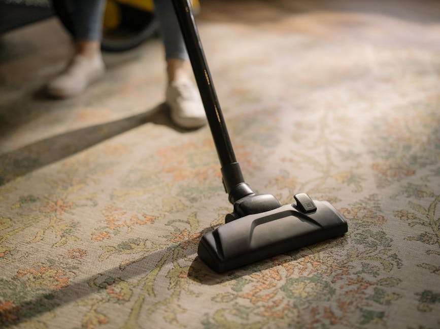 A close-up image of a vacuum cleaner with a black and grey body, actively being used to clean a patterned carpet in a room. The carpet features a floral design with light green, beige, and orange tone