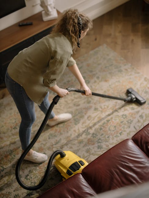 A close-up image of a vacuum cleaner with a black and grey body, actively being used to clean a patterned carpet in a room. The carpet features a floral design with light green, beige, and orange tone